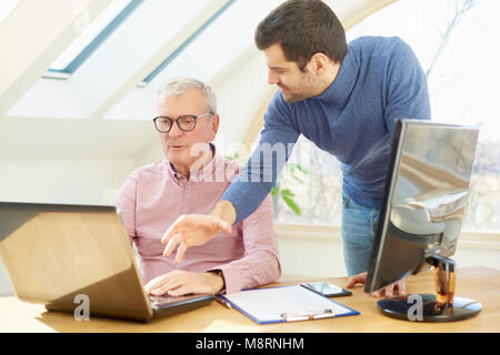 Senior businessman sitting at desk in office avant de l'ordinateur tandis que son jeune assistant businessman debout à côté de lui et la consultation sur les nouveaux pro Banque D'Images