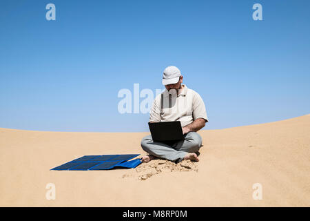 Un homme travaille sur un ordinateur portable alimenté par un panneau solaire portable dans le désert, travail à distance, autonomie et technologie écologique dans un cadre extérieur Banque D'Images