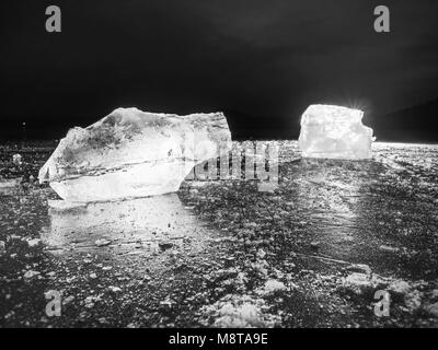 Des cubes de glace sur un terrain de réflexion sombre. Morceaux de glace haché rougeoyant et flocons de neige dans une forte contre-jour. La photographie en noir et blanc. Banque D'Images