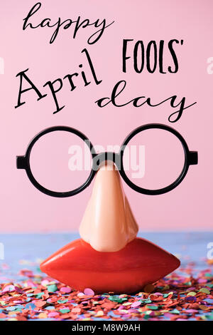 Une paire de lunettes, avec de faux nez et de la bouche, et l'happy April Fools Day, sur une surface rustique plein de confettis, contre un fond rose Banque D'Images