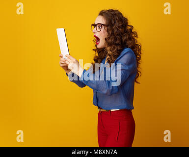 Choqué moderne femme aux longs cheveux ondulés brunette using tablet PC isolé sur le jaune Banque D'Images