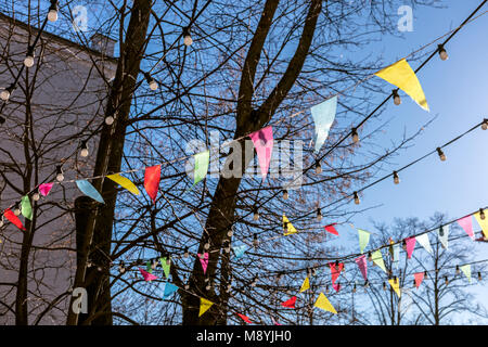 Drapeaux triangulaires colorés voletant sur les branches d'arbres contre le ciel bleu clair printemps. décoration de fête en plein air. Banque D'Images