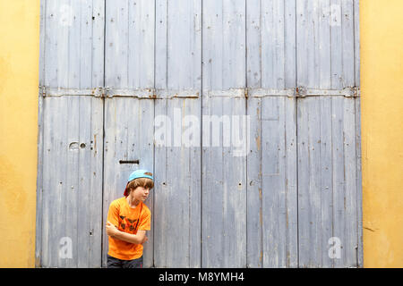 Jeune garçon avec cap en face de la vieille porte de bois, les bras croisés, scène de rue à Arles, Provence, France Banque D'Images