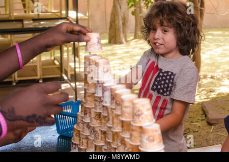 Un joli garçon aux cheveux long sourire alors qu'il travaille avec une tasse de papier sur la construction de la tour. Les mains de brown-skinned girl peut être vue de l'aider. Banque D'Images