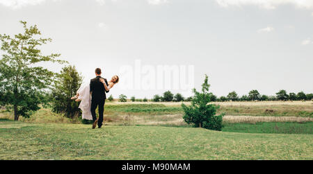 Beautiful wedding couple avec groom soulevant sa mariée au parc. Vue arrière du marié portant la mariée au parc. Banque D'Images