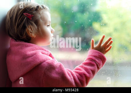 Gros plan d'une petite fille mignonne en rose peignoir regarde tristement par la fenêtre. Assis sur le rebord de la fenêtre. Banque D'Images