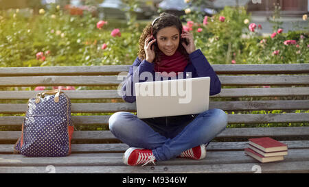 Smiling student in headphones assis sur un banc, à l'écoute de lecture des favoris. Banque D'Images