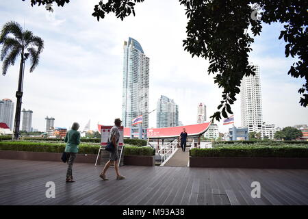 Belle photo de 2 touristes marchant sur une terrasse en bois avant l'embarquement sur un bateau avec des gratte-ciel en arrière-plan et les arbres à l'avant. Banque D'Images