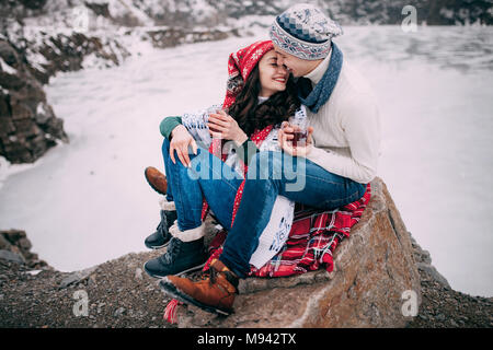 Jeune couple en chapeaux et écharpes avec tasses de thé chaud dans leurs mains est assis à côté de sourire et rock lac gelé en hiver à pied. Banque D'Images