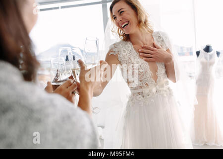 Cheerful young woman in robe de mariage toasting champagne avec des amis à bridal Boutique. Belle Mariée en robe de mariage élégante de clinking glasses Banque D'Images