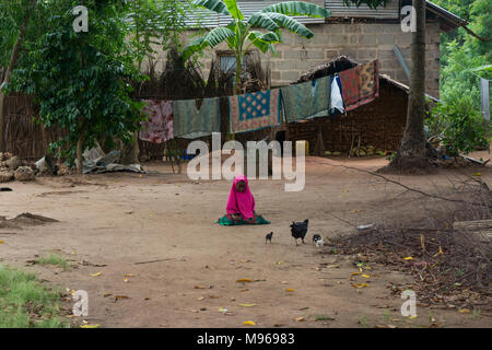 Jeune fille africaine à lonely assis dans une cour de terre en face de la maison familiale avec des poules qui courent partout. Banque D'Images