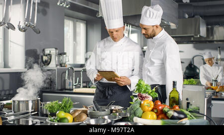 Deux grands Chefs discuter de leur blog vidéo lors de l'utilisation de l'ordinateur tablette. Ils travaillent sur un grand restaurant cuisine professionnelle en acier inoxydable. Banque D'Images