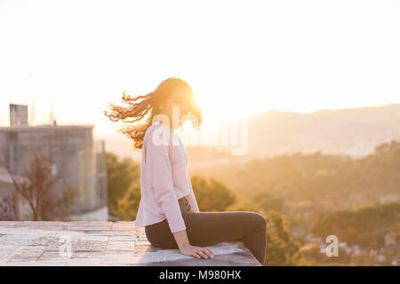 Jeune femme assise sur un mur au coucher du soleil jetant ses cheveux Banque D'Images