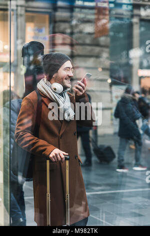 Smiling Young man with guitar case à la station using cell phone Banque D'Images