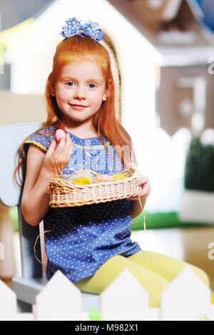 Enfant funny little Girl with red hair avec panier de Pâques sur une clôture blanche et l'arrière-plan une poupée toy house. Préparation pour la célébration de Pâques Banque D'Images