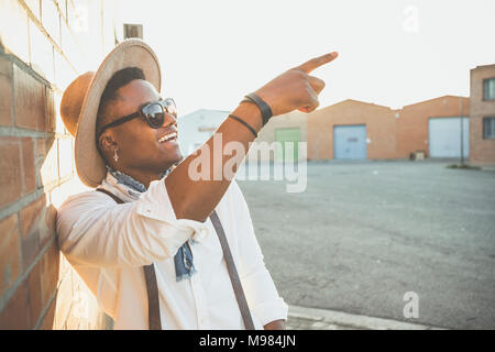 Portrait of laughing young man wearing hat and sunglasses pointant sur quelque chose Banque D'Images