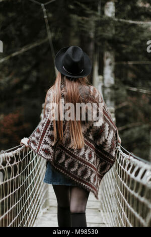 Vue arrière de la mode jeune femme portant chapeau et poncho marche sur suspension bridge Banque D'Images