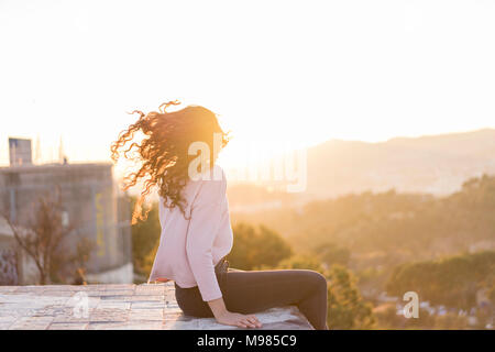 Jeune femme assise sur un mur au coucher du soleil jetant ses cheveux Banque D'Images