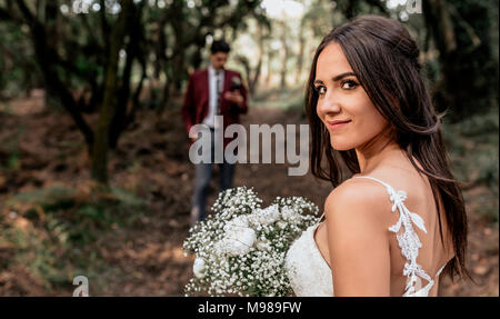 Portrait of smiling bride holding bouquet de fleurs en forêt avec le marié en arrière-plan Banque D'Images