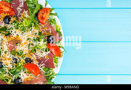 Close up de salade appétissante avec viande séchée, jambon, fromage râpé, les olives et mélanger des feuilles de laitue en plaque blanche sur table en bois bleu Banque D'Images