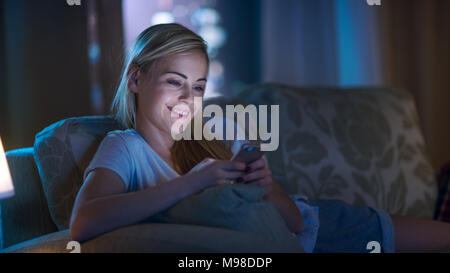 Tard dans la nuit belle femme couchée sur le canapé dans le salon, en utilisant son Smartphone et souriant. Son appartement a grande ville vue de la fenêtre. Banque D'Images