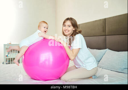 Mère avec le bébé sur la balle Pilates sur le lit dans la chambre à coucher. Banque D'Images