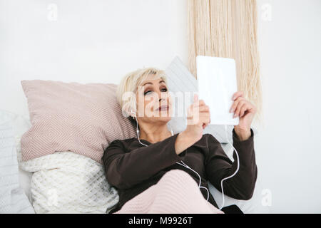 Une vieille femme positive utilise une tablette pour regarder des vidéos, écouter de la musique et discuter avec vos amis sur les réseaux sociaux. Banque D'Images