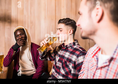 Trois jeunes hommes en vêtements décontractés sont souriants de boire une bière tout en restant assis à comptoir de pub Banque D'Images