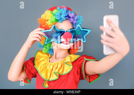 Fille de bande dessinée dans un costume de clown et stupides lunettes holding smart phone selfies faire photo. Studio shot, isolé sur fond gris Banque D'Images