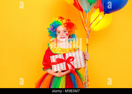 Concept d'anniversaire. Petit clown holding beaucoup de ballons et boîte-cadeau . Studio shot, isolé sur fond jaune Banque D'Images