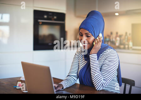 Jeune femme entrepreneur arabe portant un hijab assis à sa table de cuisine à la maison l'utilisation d'un cellulaire et à l'aide d'un ordinateur portable Banque D'Images