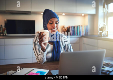 Jeune femme entrepreneur arabe portant un hijab travaillant sur un ordinateur portable et de boire du café tout en étant assis à une table dans sa cuisine Banque D'Images