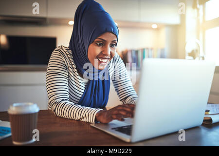 Souriante jeune femme arabe portant un hijab assis à sa table de cuisine à l'aide d'un ordinateur portable tout en travaillant de chez vous. Banque D'Images