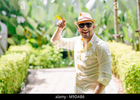Jeune homme dans le port de lunettes de soleil chapeau jaune et les tenues de s'amuser dans les jours ensoleillés. Contexte Tropical. Banque D'Images