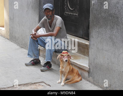 Un homme est assis sur un pas à côté d'un chien portant des lunettes et un chapeau de paille dans une rue de la Vieille Havane, Cuba. Banque D'Images