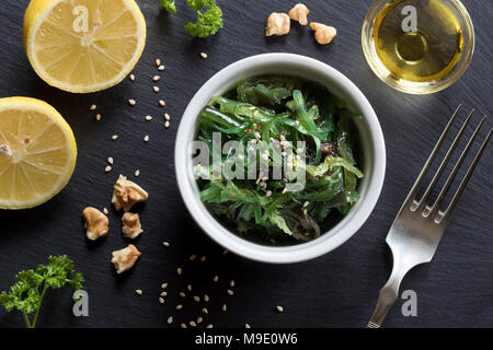 Salade faite d'algues fraîches dans un bol sur une table, avec du citron, l'huile, le persil, les noix et les graines de sésame dans l'arrière-plan, Vue de dessus Banque D'Images