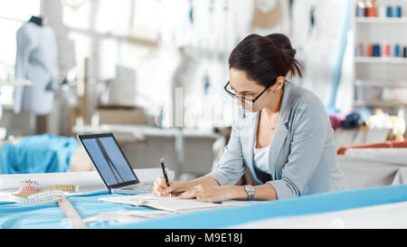 Beau la mode, designer, est assis à son bureau, utilise le smartphone. Son studio est ensoleillée, pleine de détails colorés, tissus, articles de couture, de croquis. Banque D'Images