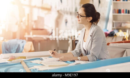 Belle femme la mode, designer, est assis à son bureau et s'inspire des croquis. Son studio est ensoleillée, pleine de détails colorés, tissus, articles de couture, Sketch Banque D'Images