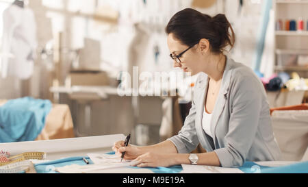 Belle femme la mode, designer, est assis à son bureau et s'inspire des croquis. Son studio est ensoleillée, pleine de détails colorés, tissus, articles de couture, Sketch Banque D'Images