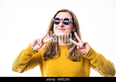 Une jeune femme de race blanche fille portant un chandail de cavalier jaune, à la cool, le port de lunettes de soleil , en donnant un double 'paix' signe avec ses doigts , sur un fond blanc, UK Banque D'Images