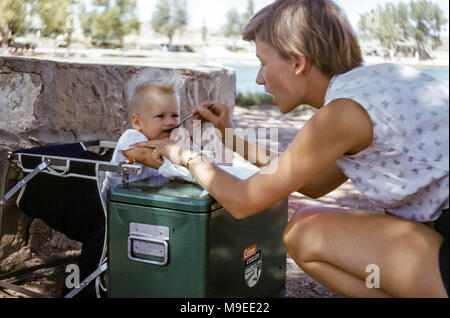 Jeune femme attrayante qui allaite un enfant de 7 à 8 mois Petite fille dans un camping aux Etats-Unis dans les années 1950 avec une grande glacière de camping en métal vert Banque D'Images