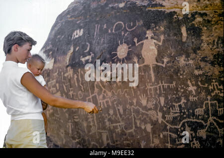 Jeune femme attrayante avec des cheveux courts tenant une petite fille de 7 à 8 mois regardant les pétroglyphes indiens indigènes sculptés sur un rocher, le journal Rock Petroglyphes, Petrified Forest National Park, Arizona, États-Unis dans les années 1950 Banque D'Images