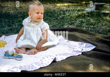 Bébé fille blonde aux cheveux de 4 à 5 mois portant une robe à smocks assise sur un tapis de pique-nique dans un jardin avec des hochets à jouets, San Jose, Californie, États-Unis dans les années 1950 Banque D'Images