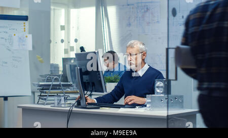 Bureau d'ingénierie d'occupation. Ingénieur travaillant à son bureau, certains éléments techniques étendu sur son bureau.Certaines personnes marchant autour. Banque D'Images