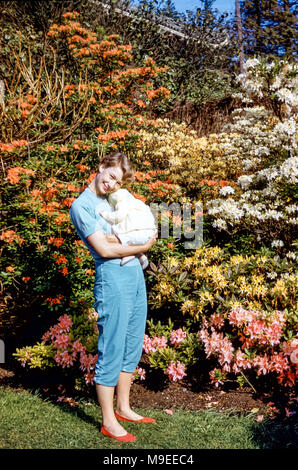 Jeune jolie femme debout devant des buissons de rhododendrons colorés, tenant un bébé endormi de trois mois, Port Defiance Park, Tacoma, État de Washington, États-Unis dans les années 1950 Banque D'Images