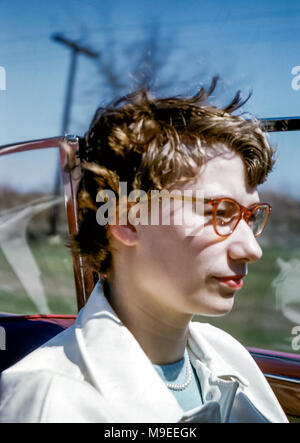 Gros plan d'une jeune femme portant des lunettes et un rouge à lèvres avec des cheveux soufflant dans le vent tout en conduisant un rouge Cabriolet Plymouth 1952 avec capote abaissée Une route aux États-Unis dans les années 1950 Banque D'Images