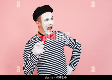 Homme dans un pullover rayé, gants blancs wink et montre de l'index. Clown, artiste , mime. Fond rose, Studio shot Banque D'Images