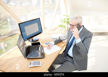 High angle shot de consultant financier senior businessman wearing suit alors qu'il était assis à un bureau en face de l'ordinateur portable et faire appel. Banque D'Images