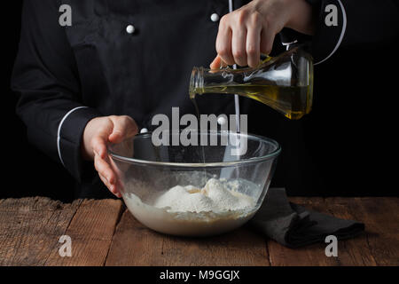 Les mains du chef de l'huile d'olive coule dans le bol sur pâte crue sur un fond noir, cuisson de la pâte à pizza. Banque D'Images