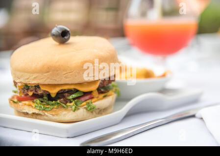 Un tir de cheeseburger classique, surmontée d'une olive noire. Un cocktail orange vif/jus dans l'arrière-plan. Banque D'Images